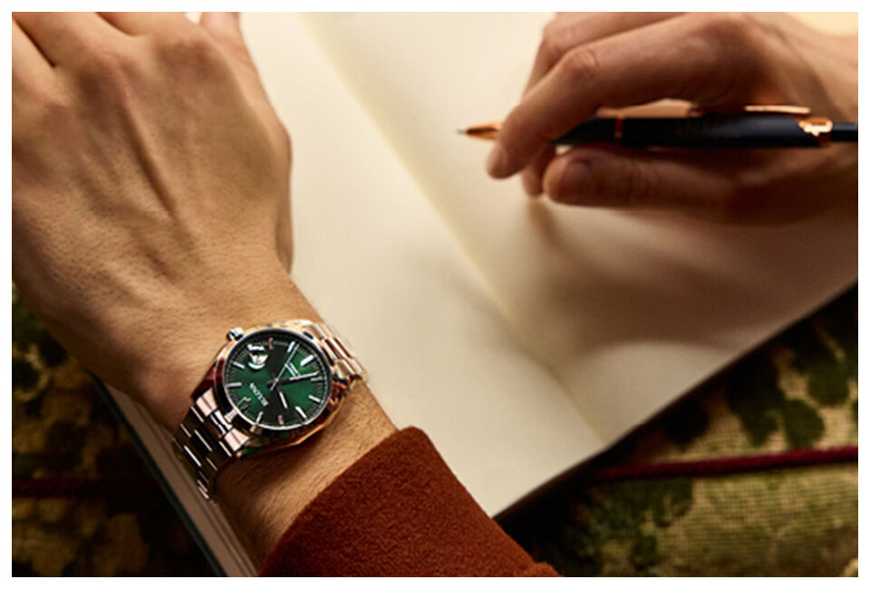 Close-up of man wearing Bulova green-dial stainless steel watch while writing in notebook.
